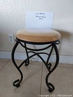 Single vanity stool with beige round cushioned seat covered in clear plastic and a black metal frame with curled decorative legs.