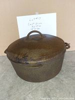 Photo showing a large cast iron kettle with handle on top of the cover. Textured surface and two side handles are visible. A handwritten note behind the kettle reads 'Lot #17 Cast Iron Kettle 12" Wide'.