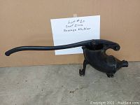 Front angled view of a black cast iron sausage stuffer with a long handle resting on a concrete floor, accompanied by a handwritten sign indicating 'Lot #20 Cast Iron Sausage Stuffer'.