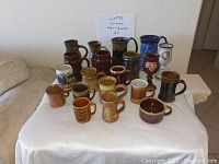 Photo showing full set of 20 ceramic mugs and goblets in various colors and sizes arranged side by side on white table covering