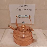 Photo of solid copper tea kettle with handwritten sign 'Lot #74 Copper Tea Kettle' in background, showing porcelain handle and lid knob with blue decorative pattern. Light dent on upper right side visible.