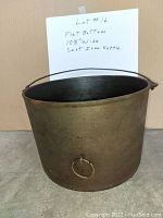 Front-facing view of a large cast iron kettle with flat bottom, showing side pouring ring and wire handle attached to the rim.