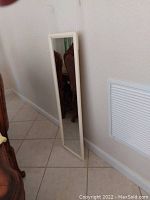 Full-length image of a rectangular mirror with white frame leaning against a white wall on tiled floor, showing reflection of wooden chair.