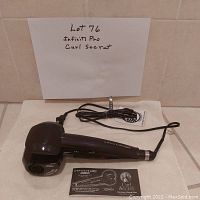 Black Infiniti Pro Curl Secret automatic hair curler placed on a flat surface with power cord coiled beside it. Instruction booklet positioned in front. Background shows a handwritten paper with lot number and item name.