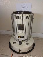White metal cylindrical kerosene heater with metal protective cage and control knobs on base, positioned on floor against a wall. Includes handwritten note indicating lot number and that heater is like new with a book.