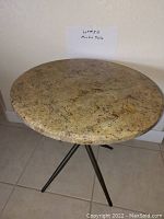 Photo showing a round marble top table with variegated beige and brown veining and four black metal legs, placed near a wall on tiled floor.