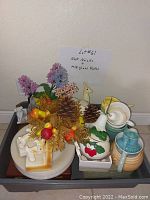 Milk glass plates stacked at bottom, various decorative ceramic and knick knacks displayed on top including artificial flowers, pine cones, small figurines, and ceramic pots