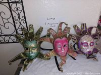 Three Venetian jester-style masquerade masks displayed on a marble surface with a wire decorative panel in the background.