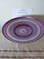 Photo showing a round decorative plate with purple and silver concentric pattern design, placed on a beige fabric.