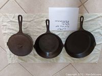 Three cast iron cookware pieces laid flat on a cloth: an 8-inch kettle with cover on the left and two fry pans of 9 and 10 inches to the right.