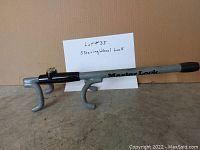 Photo of a silver and black Master Lock steering wheel lock with 2 hooks and black handle grip, includes 2 keys and lot identification card.