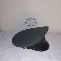 Grey U.S. Army dress uniform cap size 6 3/4 with black visor and chin strap, gold side buttons, displayed against a light wall with a handwritten note indicating lot number and item description.