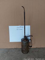 Front view of the tall vintage metal oil can with long narrow spout and handle. Note the replaced plastic cap on the container top and signs of rust and age on the body.