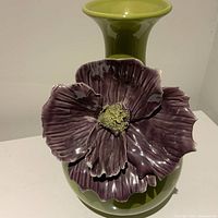 Front view of green ceramic vase with large attached purple flower showing flower detail and vase shape.