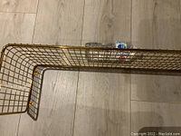 Top-down view of curved gold wire shelf laying flat on light wood floor, showing the grid pattern and right angled ends, with a water bottle for scale.