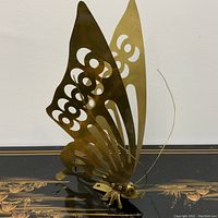 Brass butterfly sculpture photographed against a white wall and black surface to show details of wing cut-outs and antennae.