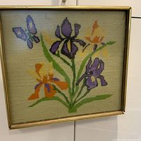 Square framed needlepoint artwork depicting purple and orange irises with green stems and a butterfly. Needlepoint shows pixelated floral design on a beige background. Frame is gold-toned and shows slight wear.