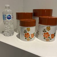 Four vintage metal kitchen canisters with matching brown lids and retro floral designs, displayed next to a water bottle for scale.