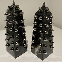 Two black Carrara marble obelisks with silver metal spikes shown side by side from front right angle, illustrating spike design and marble veining.