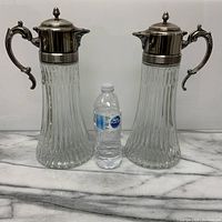 Front view of pair of large matched glass claret jugs with silver tops and handles, shown next to a water bottle for size comparison.