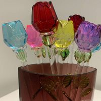 Close-up of eight colorful crystal rose swizzle sticks arranged inside the decorative red glass vase, showing the rose buds and green petals.