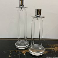 Two clear lucite candlesticks with metal candle holders on a black surface against white background, showing full height and shape.