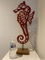 Front view of large red cast metal sea horse sculpture on wooden base next to a water bottle for size scale.