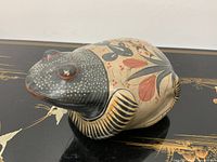 Side angle view showing the black head with white spots and orange-rimmed eyes, beige body with floral patterns, and front legs striped in black and orange.