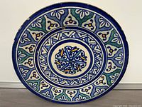Full front view of the round hand-painted plate showing the intricate floral and geometric design in blue, green, yellow, and white.