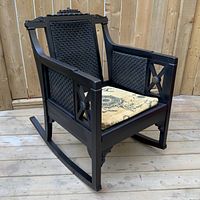 Angled full view showing black painted frame, carved top rail, and woven bamboo caning