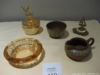 Full group shot showing all items: rose glass ashtray, brass ashtray with sailboat figurine, pottery jug and bowl, amber glass trinket jar with deer lid.