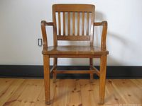 Front view of the vintage banker's chair showing solid oak construction, slatted backrest, and the left arm slightly damaged.