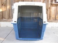 Front view of plastic travel dog crate with blue base and white top, metal wire door closed.