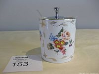 Front view of the vintage Staffordshire ceramic jam jar with floral pattern and silver lid on top with glass spoon inside.