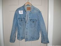 Front view of light blue denim Levi's jean jacket hanging on wooden hanger, showing overall shape and button front.