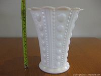 Front view of white milk glass vase next to measuring tape showing 7 inches height with hobnail bead vertical pattern and scalloped rim.