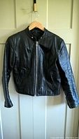 Front view of black leather motorcycle jacket on hanger, shows zipper and snap-button collar closure.