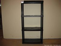Photo of a black resin shelving unit showing all four shelves and vertical support poles, against a plain beige wall and brown floor.