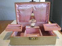 Vintage Mele wind-up music jewellery box open showing pink velvet interior, storage compartments, and dancing ballerina figure on platform. The underside of the lid has pink satin with gold-trimmed curtain design and a mirror in the center.