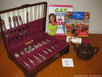 Photo of Oneida Tudor silverware set displayed in open wooden velvet-lined box along with two cookbooks, new baking mat, and Brown Betty teapot on wooden table