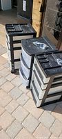 Three black framed plastic storage bins each with four white translucent drawers and black organizational tops with compartments. The wheels for each bin are packaged on top in plastic bags.