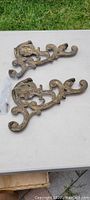 Pair of ornate antique brass shelf brackets shown on white surface, highlighting aged patina and decorative scrollwork.