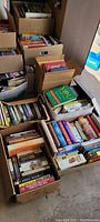 Boxes filled with a variety of books including cookbooks and fiction, stacked in a room.