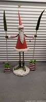 Front view of large standing Santa figure with long red hat, holding tall Christmas tree ski poles with wrapped gift bases.
