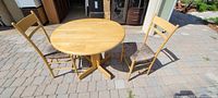 Photo showing round wood veneer drop leaf table and two light wood chairs with rush seats on outdoor stone surface.
