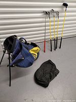 Three golf clubs with yellow shafts and one red golf club standing next to a blue and yellow golf bag with black accessory bag on the floor