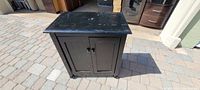 Black wood storage cabinet with two paneled front doors closed, showing short legs and flat top with surface marks.