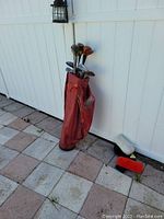 Red golf bag holding 15 assorted golf clubs outdoors against white fence. Clubs include driver and irons with steel shafts. One red head cover visible on ground.