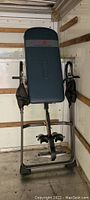 Full view of Ironman gravity inversion table upright against wall with padded blue backrest, foam covered handles, ankle holders, and tubular chrome frame, left handle foam covering partially missing.
