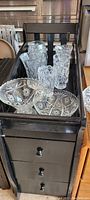 Photo showing 8 glass items including vases and bowls with various pressed glass patterns in clear glass, arranged on a wooden cart.